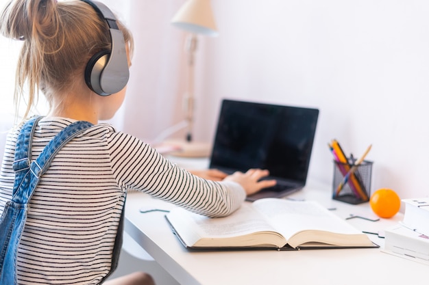 Portrait of teenage girl learning online with headphones and laptop taking notes in a notebook sitting at her desk at home doing homework