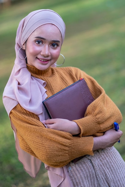 Photo portrait of teenage girl wearing headscarf holding book in park
