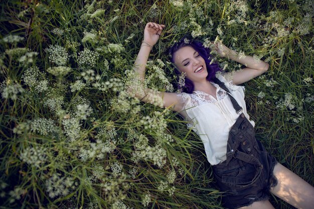 보라색 머리와 귀에 귀걸이를 한 10대 소녀의 초상화는 자연의 풀밭에 누워 있습니다.