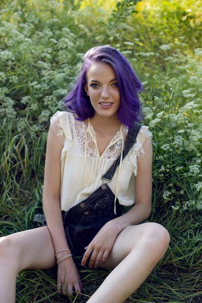Portrait of a teenage girl with purple hair and an earring in her nose sit in the grass in nature