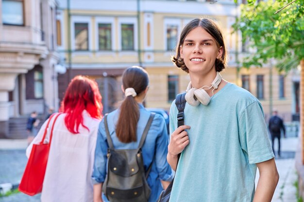 外でカメラを見ている十代の男の肖像画 笑顔の若いハンサムな男性学生 17 18歳 バックパックヘッドフォンで街の通りで