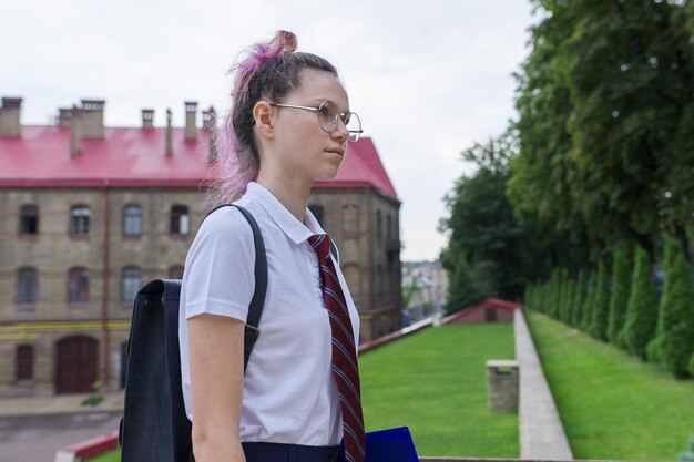 Portrait of teenager girl with backpack going to school, summer autumn morning, school building background. Back to school, back to college