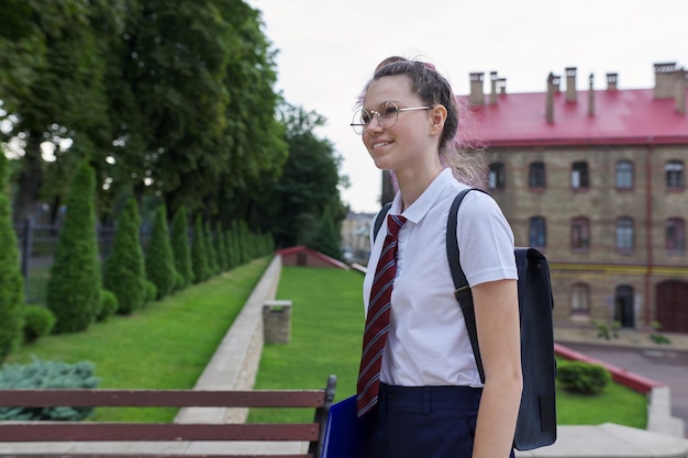 Портрет девочки-подростка с рюкзаком, идущим в школу, летним осенним утром, школьным зданием. Снова в школу, обратно в колледж
