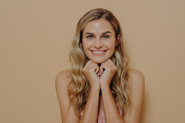 Portrait of tender charming european female holding her hands under her chin, woman with perfect styled hair wearing summer light dress looking at camera with beaming smile and happy expression