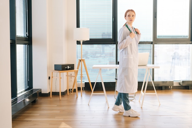 Portrait of thinking female doctor in white coat standing on background of window in sunny day in light medical clinic office.