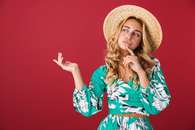 Portrait of thinking thoughtful cute young blonde girl in bright blue dress posing isolated over red wall looking aside wearing hat showing copyspace