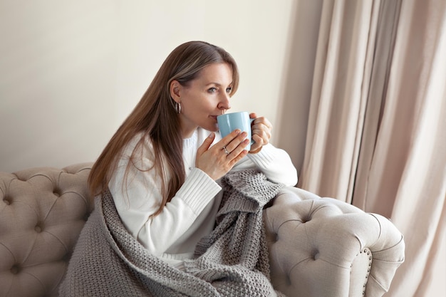 Foto ritratto pensando donna seduta a casa in un soggiorno minimalista beige sul divano, bevendo tè dalla tazza blu, vestito con plaid lavorato a maglia, cosy hygge. modello caucasico con capelli lunghi che indossa un maglione caldo