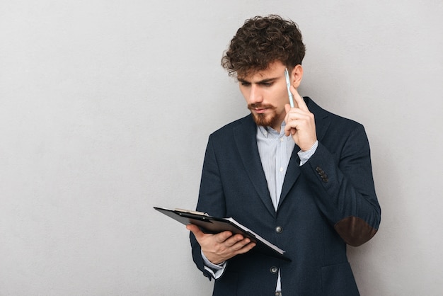 portrait of a thinking young business man isolated over grey wall holding clipboard.