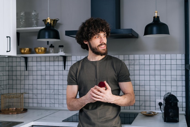 Портрет думающего молодого человека, держащего коробку с обручальным кольцом и мечтающего смотреть в окно на кухне дома