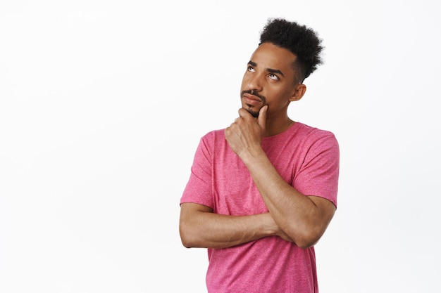 Portrait of thoughtful african american guy rub chin, looking up and thinking, ponder decision, making choice, standing in pink t-shirt against white background. Copy space