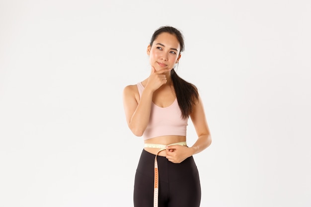 Photo portrait of thoughtful asian female gym coach, sportswoman thinking about losing more weight, measuring waist after workout, white background.