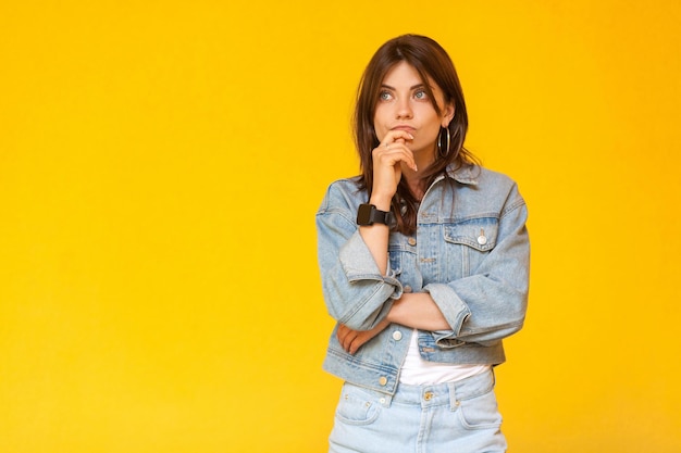 Portrait of thoughtful beautiful brunette young woman with makeup in denim casual style standing, touching her lips, looking away and thinking what to do. studio shot, isolated on yellow background.