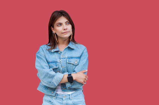 Portrait of thoughtful beautiful brunette young woman with makeup in denim casual style standing with crossed hands and looking away thinking. indoor studio shot, isolated on red background.