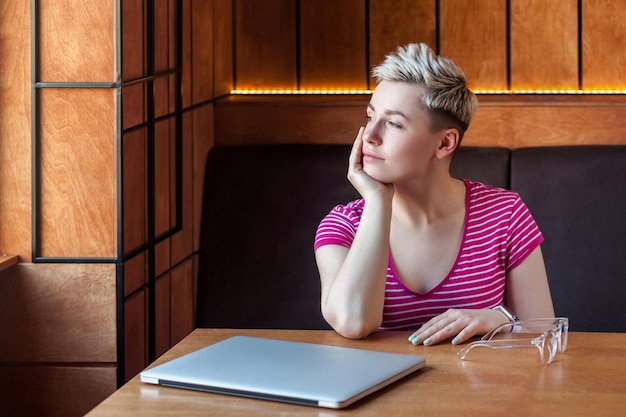 Photo portrait of thoughtful beautiful young bussineswoman with blonde short hair in pink tshirt is sitting in cafe finishing work and looking away relaxing and smile indoor healthy lifestyle