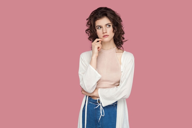 Portrait of thoughtful brunette young woman with curly hairstyle in casual style standing, touching her chin, looking away and thinking what to do. indoor studio shot isolated on pink background.