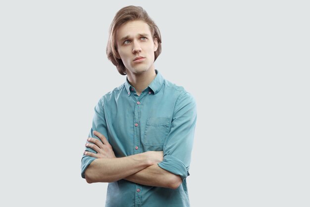 Portrait of thoughtful or confused handsome long haired blonde man in blue casual shirt standing with crossed arms, looking away and thinking. indoor studio shot, isolated on light grey background.