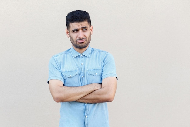 Portrait of thoughtful confused handsome young bearded man in blue shirt standing, crossed arms, looking away and thinking what to do. indoor studio shot isolated on light beige wall background.