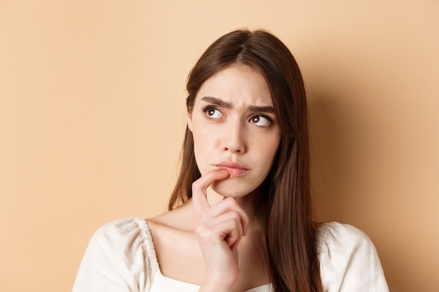 Portrait of thoughtful girl frowning and looking aside pensive, thinking or making choice, standing on beige background.