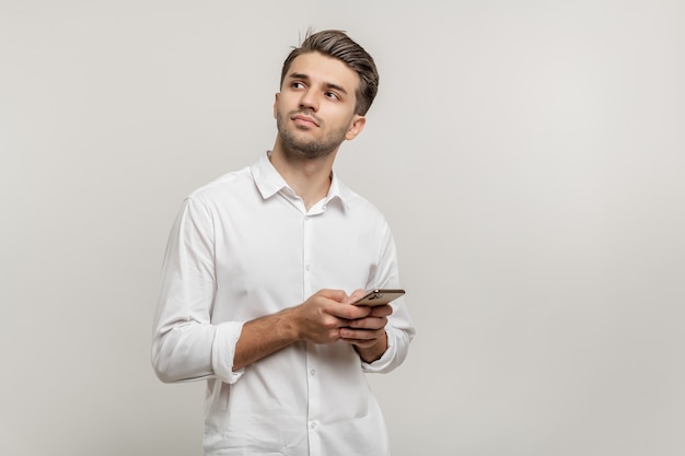 Portrait of thoughtful guy wearing white shirt holding cellphone isolated against white background