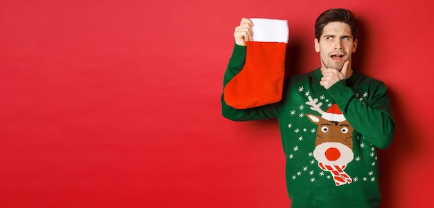 Portrait of thoughtful handsome man in green sweater holding christmas stocking and thinking guessin...