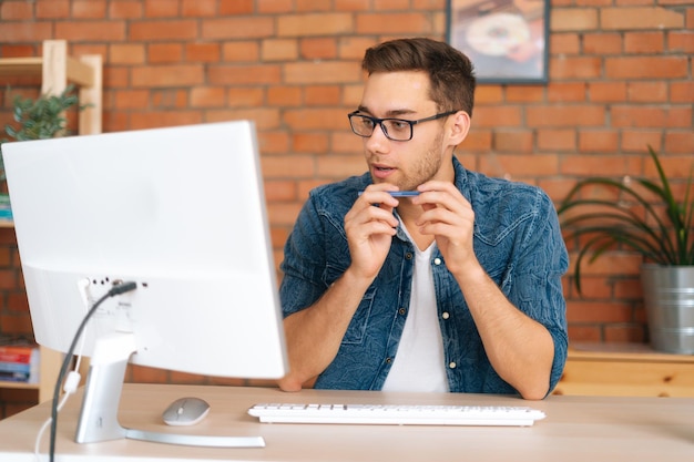 Portrait of thoughtful handsome young freelance programmer male in stylish glasses working on desktop computer holding pen in hands