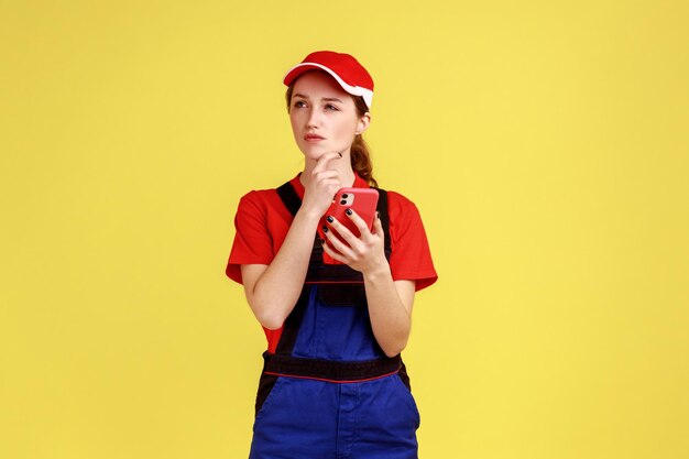 Portrait of thoughtful handy woman using mobile phone for online supporting having pensive expression holding chin and looking away Indoor studio shot isolated on yellow background