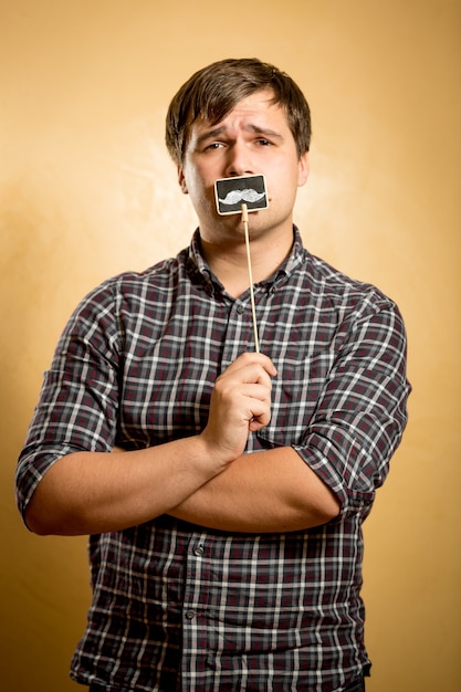 Portrait of thoughtful hipster guy posing with fake mustache on stick