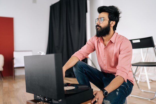 Portrait of thoughtful Indian hipster in glasses listening to music on old record player vinyl discs sitting on floor Music lover man listening to classical music