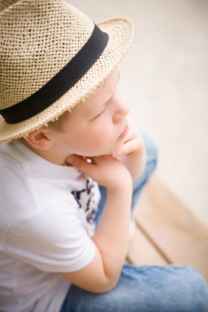 Portrait of a thoughtful kid boy in a straw hat sitting on a wooden bench and looking into distance