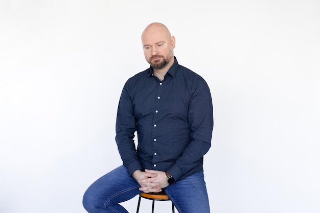 Portrait of thoughtful middleaged man in blue shirt jeans\
sitting on stool interlacing fingers on wh