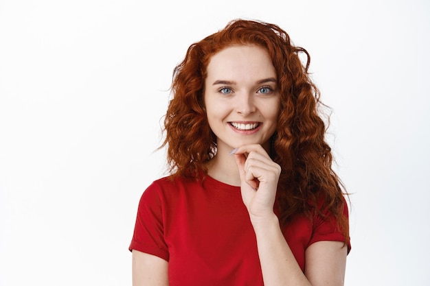 Foto ritratto di modella rossa premurosa con trucco di luce naturale, toccando il mento e guardando pensieroso, pensando a qualcosa, muro bianco