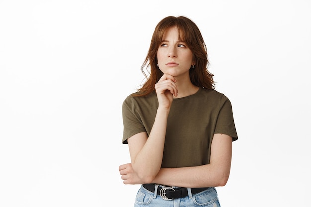 Portrait of thoughtful redhead woman looking at upper left corner, touch chin and thinking, pondering or making choice, deciding, standing on white