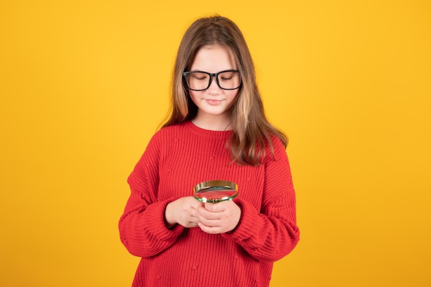 Portrait of thoughtful teenage girl in eyeglasses holding magnifying glass and looking Teen girl on yellow surface