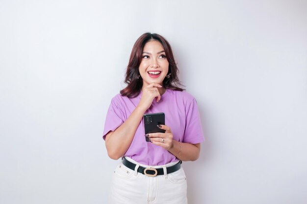 Портрет задумчивой молодой азиатки в сиренево-фиолетовой футболке, смотрящей в сторону и держащей смартфон