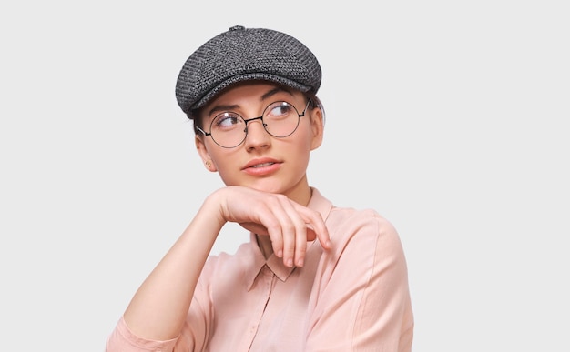 Портрет вдумчивой молодой женщины в прозрачных очках, повседневной розовой рубашке и серой кепке, думающей и серьезно смотрящей на пустое место для копирования вашей рекламы или рекламной информации