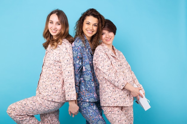 Portrait of three beautiful young girls wearing colorful pyjamas having fun during sleepover isolated over blue background. Pajama party and hen-party concept