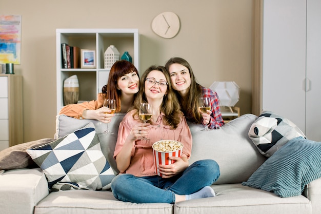 Портрет трех веселых молодых подружек с мисками для попкорна и вином, возле стильного дивана дома.