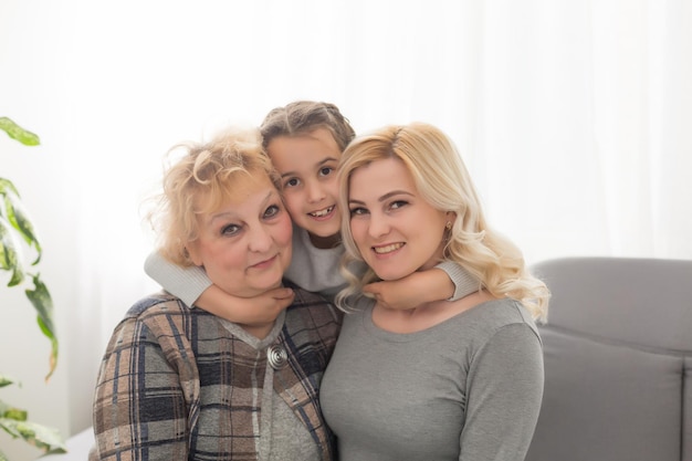 Portrait of three generations of women look at camera posing for family picture, cute little girl hug mom and granny enjoy time at home, smiling mother, daughter and grandmother spend weekend together