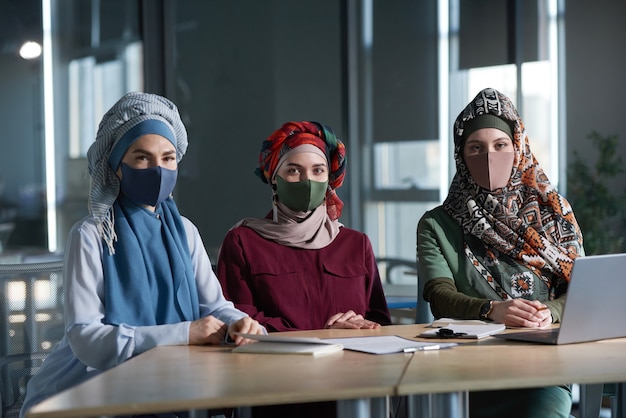 사무실에서 함께 일하는 동안 카메라를 바라보는 전통 의상과 보호 마스크를 쓴 세 명의 이슬람 여성 초상화