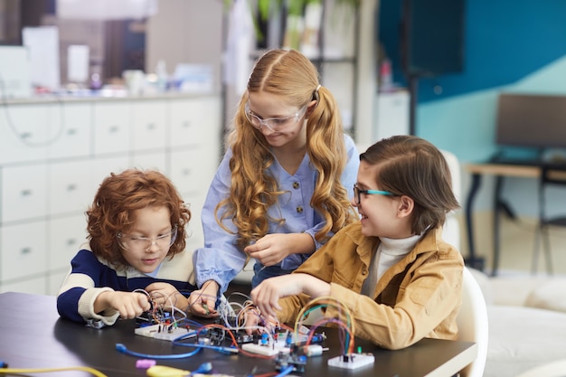 Портрет трех улыбающихся детей, экспериментирующих с электрическими цепями во время сборки роботов в классе инженеров в школе