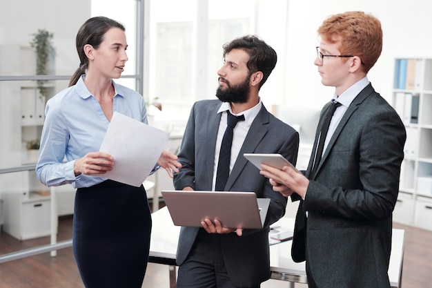 Portrait of three young business people discussing work and smiling while standing in modern office