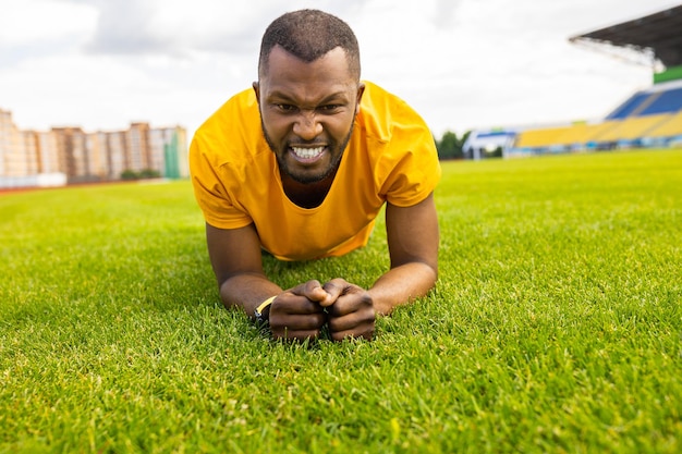 Portrait of a tired african american male working out hard on a grass field at the stadium Young athletic man doing plank exercise outdoors in yellow sportswear training concept