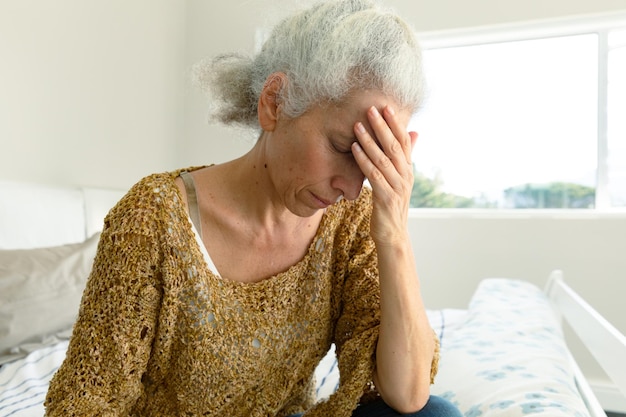 portrait of tired caucasian senior woman with headache sitting on bed at home