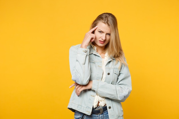 Portrait of tired dissatisfied young woman in denim casual\
clothes looking aside, putting hand on head isolated on yellow\
orange background in studio. people lifestyle concept. mock up copy\
space.