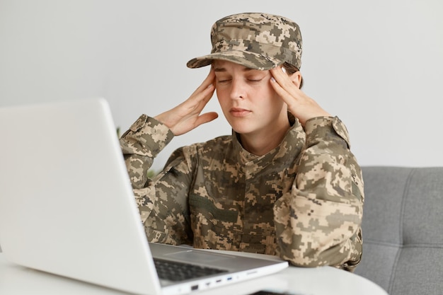 Portrait of tired exhausted woman soldier wearing camouflage uniform and hat, posing at home, sitting in front of laptop computer and touching her head, suffering headache, being tired.