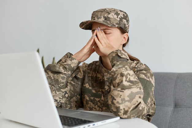 Portrait of tired female soldier wearing camouflage uniform and hat sitting in front of laptop computer and touching her eyes suffering headache being tired working long hours