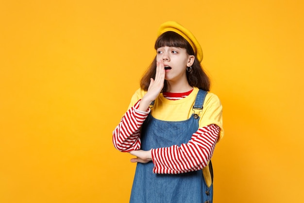 Portrait of tired girl teenager in french beret, denim sundress yawning, covering mouth with hand isolated on yellow wall background. people sincere emotions, lifestyle concept. mock up copy space