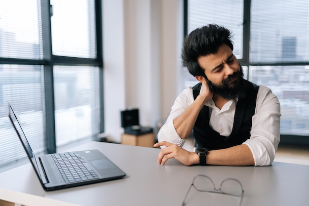 Portrait of tired Indian businessman in stylish clothes suffering from fibromyalgia neck pain working on laptop sitting at office desk by window