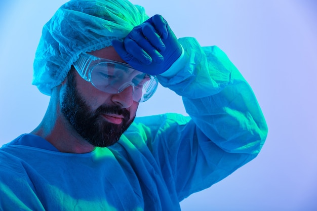 Portrait of a tired man scientist wearing unifrom and glasses standing isolated over blue-pink haze , wiping forehead