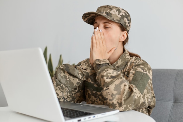 Portrait of tired military woman wearing camouflage uniform and\
cap sitting and working on laptop having exhausted look keeps eyes\
closed trying to rest